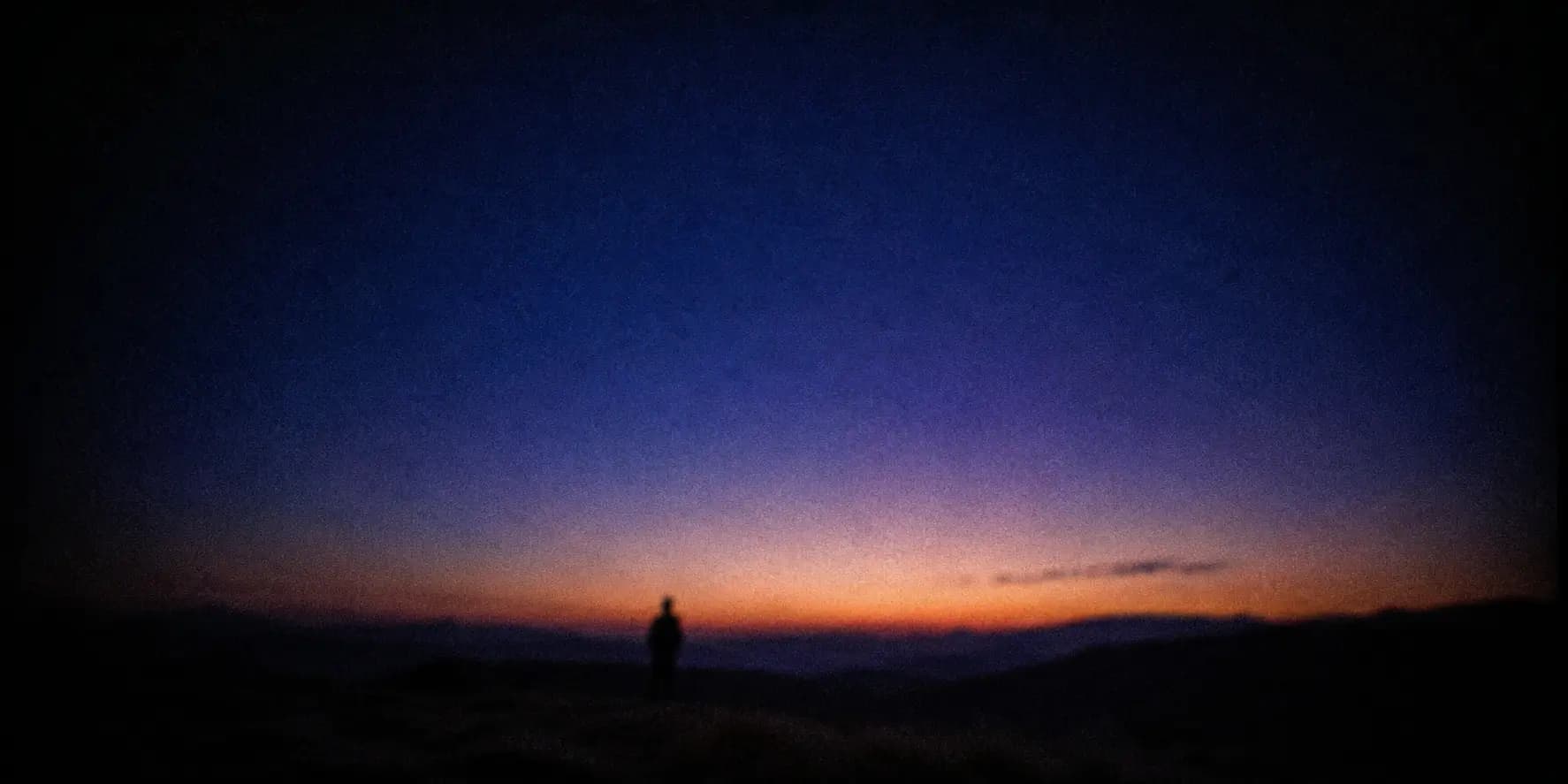 A lone figure on a hillside at dusk, watching the day fade from a warm amber horizon up into deep cool indigo